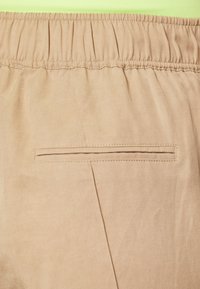 Beige bukse med elastisk midje og en enkelt baklomme, med subtil tekstur. Har et rent, jevnt design med detaljerte sømmer.