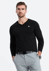 Maglietta nera a maniche lunghe con scollo a V e logo. Abbinata a pantaloni grigi chiari e una cintura nera. Orologio argento visibile sul polso.