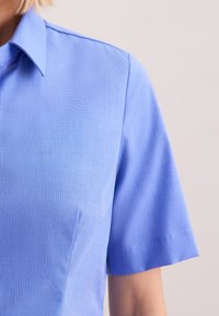Blusa a maniche corte in tessuto azzurro chiaro, con finitura testurizzata, colletto a punta e dettagli di cucitura sottili sulle maniche.