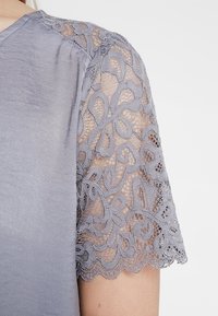 Blusa de manga corta en tela de color gris claro, con detalle de encaje en los hombros con patrones florales y bordes ondulados.