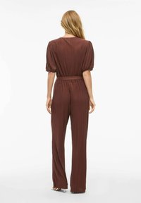 Ruda pilota suknelė su trumpomis, pūkuotomis rankovėmis, dirželiu liemeniui ir plačiomis kelnėmis, pasižyminti lygia tekstūra ir subtiliu spindesiu.