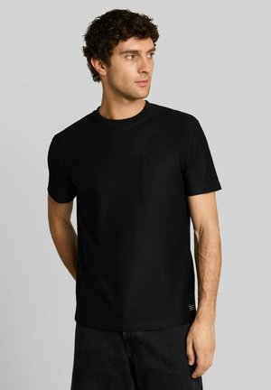 Mann trägt ein schlichtes schwarzes T-Shirt mit kurzen Ärmeln und Brusttasche, schaut nach links und steht vor einem einfarbigen hellgrauen Hintergrund.