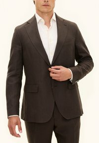 Giacca da completo in lino marrone con rever a punta, due tasche frontali e polsini con bottone. Indossata su una camicia di colore chiaro. Texture liscia.