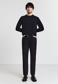Giovane uomo con capelli ricci scuri, che indossa un maglione nero, pantaloni neri con cintura e scarpe lucide nere, in piedi su uno sfondo chiaro e uniforme.