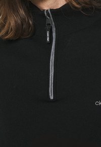 Schwarzer Reißverschluss-Pullover mit hohem Kragen, ausgestattet mit einem grauen Reißverschluss und "Calvin Klein"-Markierung in Weiß auf der Vorderseite. Strukturiertes Material.