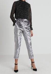 Schwarze transparente Bluse mit weißem Muster, hoher Kragen, lange Ärmel, kombiniert mit silbernen, metallischen, knöchellangen Hosen mit strukturiertem Finish. Schwarze Absätze.
