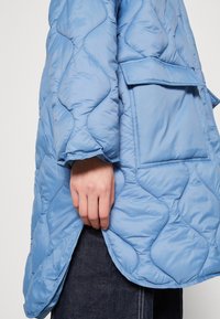 Lyseblå quiltet jakke med en glat overflade, der har en laplomme og en rundet kant, vist iført over mørk denim.