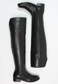Une paire de bottes en cuir noires, montantes jusqu'au genou, l'une debout et l'autre montrant la fermeture éclair intérieure, avec des talons blocs bas sur fond blanc.