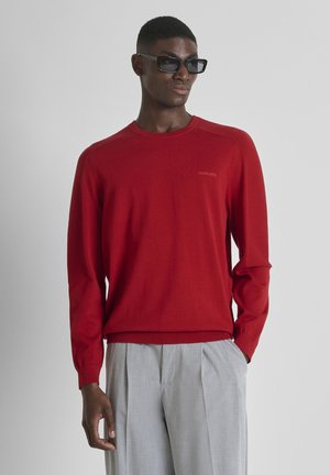 Homme portant des lunettes de soleil noires, un pull rouge à manches longues avec des manches texturées, et un pantalon plissé gris clair, debout devant un fond uni.