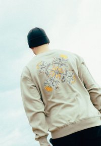 Sweatshirt beige clair avec un motif de broderie florale jaune et gris au dos. Le modèle porte un bonnet noir. Texture lisse.