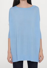 Femme portant un pull bleu clair oversize avec un large col et des manches trois-quarts, associé à un pantalon foncé.