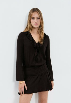 Joven con cabello largo y rubio, vistiendo un vestido negro de manga larga con cuello fruncido y una pulsera dorada, de pie contra una pared blanca lisa.