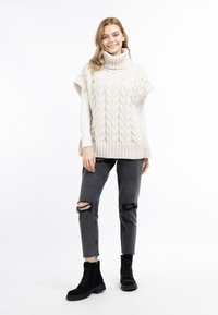 Beige gestrickter Oversize-Pullover mit hohem Kragen, der ein Zopfmuster aufweist. Kombiniert mit ausgefransten schwarzen Jeans und geschnürten schwarzen Stiefeln.