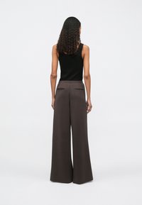 Frau mit lockigem Haar, die ein schwarzes ärmelloses Oberteil und weit geschnittene dunkelbraune Hose trägt, steht mit dem Gesicht zur Wand vor einem schlichten Hintergrund.