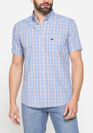 Uomo che indossa una camicia azzurro chiaro a maniche corte con bottoni, con motivo a quadri rosa e blu navy, e jeans azzurri chiari.