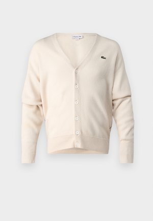 Béžový cardigan vyrobený z mäkkej látky, s výstrihom do V, zapínaním na gombíky, dlhými rukávmi a malým zeleným logom na ľavej hrudi.