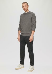 Grijze gebreide trui met geribbelde manchetten, gecombineerd met zwarte broek met streepjes en lichtgekleurde sneakers. Model staat tegen een neutrale achtergrond.