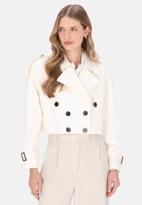 Frau mit welligem blondem Haar trägt eine kurz geschnittene, doppelt geknöpfte weiße Jacke mit großen schwarzen Knöpfen und beigen, hoch taillierten Hosen.