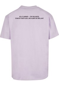 Korte mouwen lila T-shirt met een ronde hals. Zwarte tekst op de achterkant zegt: "WEES EEN FLAMINGO – BLIJF IN BALANS..."