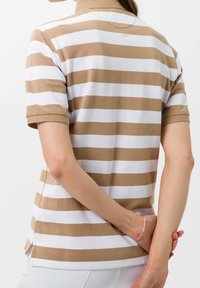 Polo tričko pruhované v béžové a bílé barvě, s krátkým rukávem, límečkem a bočními rozparky. Tkanina vypadá jemně a mírně texturovaně.