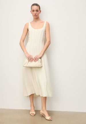 Femme portant une robe midi crème sans manches tenant une pochette assortie en tissu doux, debout devant un fond clair uni.