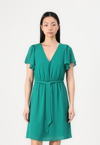 Robe verte avec un décolleté en V, des manches courtes et flottantes, et une taille élastique. Tissu lisse et léger qui tombe doucement. Ceinturée à la taille.