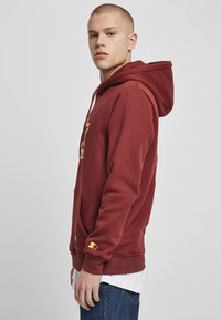 Sudadera burdeos con un bolsillo canguro, logo dorado en el pecho, capucha ajustable, puños acanalados y dobladillo. Combinada con vaqueros azules.
