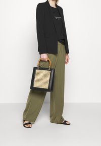 Žena držící černou kabelku s tkaným předním panelem a bambusovými držadly, oblečená v černém blejzru, černých sandálech, olivových širokých kalhotách a černém tričku.