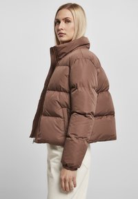 Brun pufferjakke med høj krave, der har et cropped design, polstrede sektioner og elastiske cuffs. Glat tekstur, lynlåslukning, sidelommer.