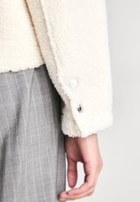 Cremfarbener Fleece-Jacke mit strukturiertem Material und silbernen Knopfdetails, getragen über grauen gestreiften Hosen.