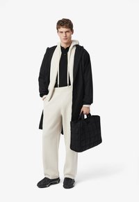 Cappotto lungo nero, felpa panna, polo nera, pantaloni sportivi panna, sneakers nere con dettagli verdi e borsa trapuntata nera.