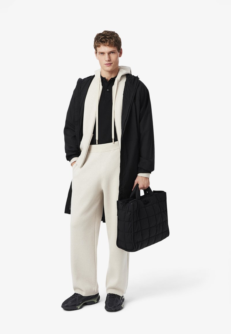 Manteau long noir, sweatshirt crème, polo noir, sweatpants crème, baskets noires avec accents verts, et un sac matelassé noir.