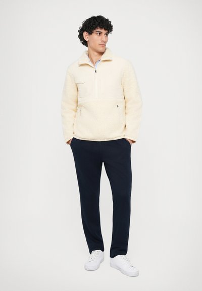 Pullover de fleece em off-white com fecho de meio-zíper, painel frontal em contraste e bolsos laterais; combinado com calças azul-escuro e ténis brancos.