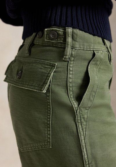 Prim-plan cu pantaloni verde măsliniu cu buzunar cu clapetă nasture și cu bretele pentru curea, purtați cu un pulover închis la culoare cu model reiat.