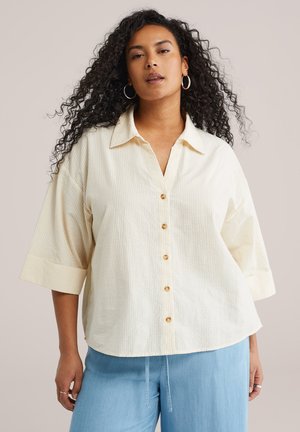 Kvinde med langt krøllet hår iført en løs cremefarvet skjorte med knapper og albuelange ærmer samt lyseblå bukser, stående foran en ensfarvet baggrund.