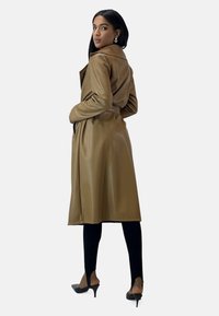 Brown faux leather trench coat with a belted waist, long sleeves, and side pockets, paired with black leggings and pointed heels.