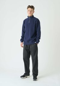 Marineblauer Fleece-Pullover mit Halbreißverschlusskragen, langen Ärmeln und gerippten Bündchen; kombiniert mit schwarzen weit geschnittenen Hosen und schwarzen Sneakern.