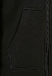 Juodas zip-up džemperis su priekinėmis kišenėmis ir tekstūrine medžiaga. Dėmesys siuvimo detalėms aplink kišenę ir užtrauktuką.