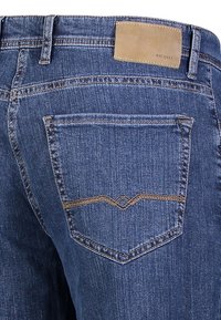 Blå denimjeans med en bakficka med ett dekorativt sömnadsmönster och en ljusbrun läderetikett med präglad text.