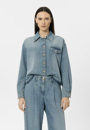 Frau mit glatten braunen Haaren, die ein hellblaues Denimhemd mit goldenen Knöpfen und eine passende hoch taillierte Denimhose trägt, stehend vor weißem Hintergrund.