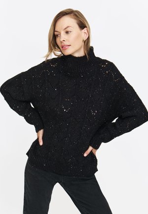 Czarny, oversize sweter dzianinowy z wysokim kołnierzem, z teksturowanym wzorem i subtelnym blaskiem w całej strukturze. Długie rękawy oraz ściągacz u dołu.