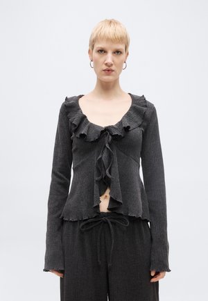 Person med kort blondt hår iført mørkegrå, tekstureret bluse med lange ærmer, flæser og bindebånd foran, parret med matchende bukser med snøre.