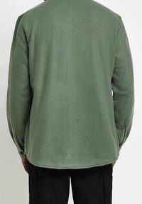 Zielona polarowa koszula z długim rękawem, zaokrąglonym dołem, mankietami na guziki i miękką fakturą. Widok z tyłu nie pokazuje żadnych wyraźnych wzorów.