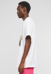 Hvid bomulds-T-shirt med korte ærmer, prydet med stor metallisk guldtekst på forsiden. T-shirten bæres med lyserøde shorts.