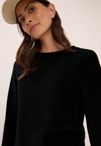 Vrouw met lang bruin haar, die een beige pet en een zwart geribde lange mouwtop draagt, kijkt iets omhoog tegen een effen achtergrond.