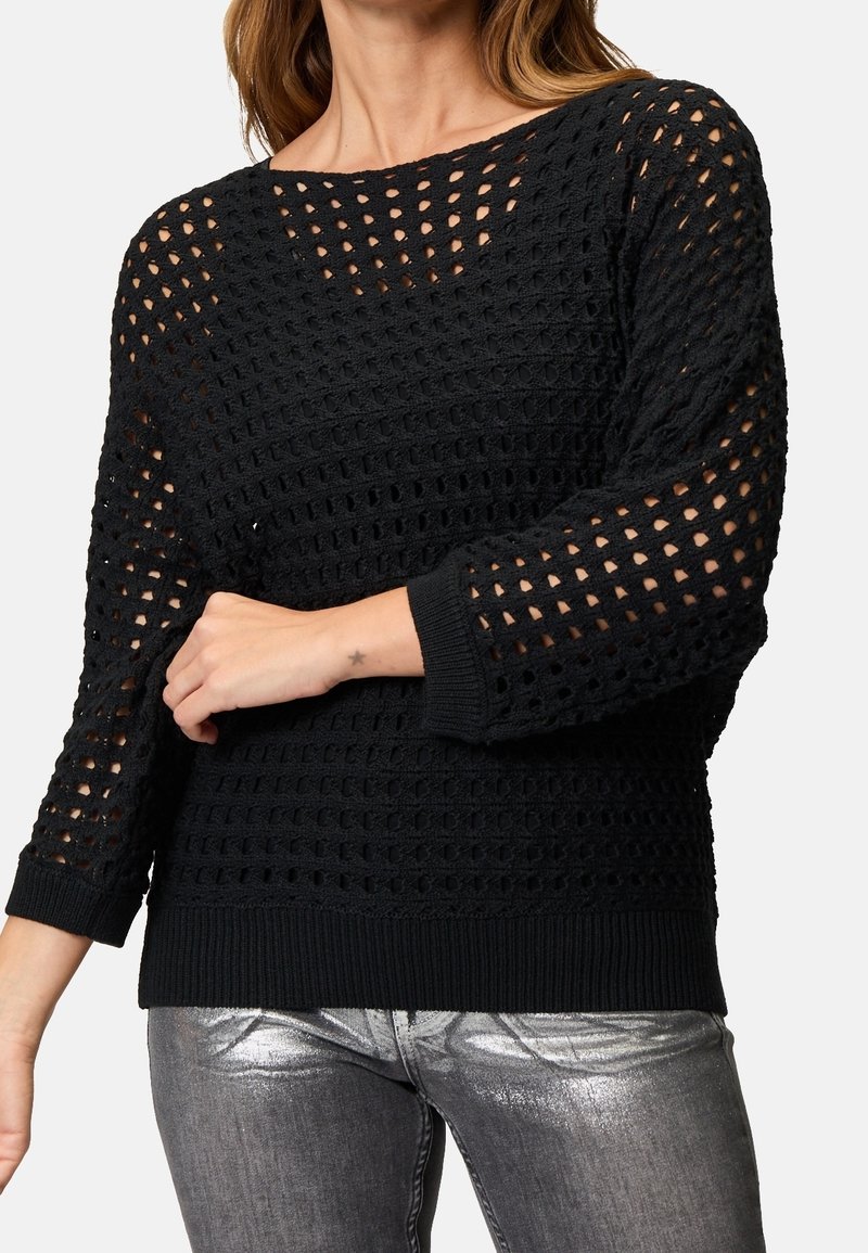 Femme portant un pull en tricot crochet noir avec des manches 3/4 et un jean argenté métallisé, montrant un petit tatouage d'étoile sur son poignet.