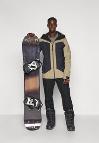 Σνόουμπορντ με μαύρη και ξύλινη υφή, κρατιέται από άτομο με μπεζ και ναυτικό μπουφάν, μαύρο παντελόνι και μαύρες χιονομποτάδες.