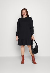 Vestido preto de mangas longas com um acabamento em babados, feito de material suave. Combinado com botas de salto castanhas e uma bolsa preta com uma alça atada.