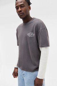 Mand iført lagdeling af grå og hvide langærmede skjorter samt lyseblå jeans, stående mod en ensfarvet hvid baggrund.