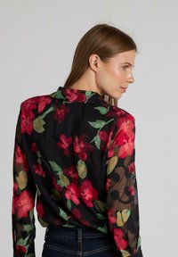 Blusa floral con una tela negra transparente, con patrones florales en rojo y verde. Diseñada con un cuello y puños abotonados.